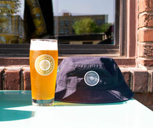 Load image into Gallery viewer, A glass of beer sits on a table next to an Indeed Bucket Hat by Legacy.
