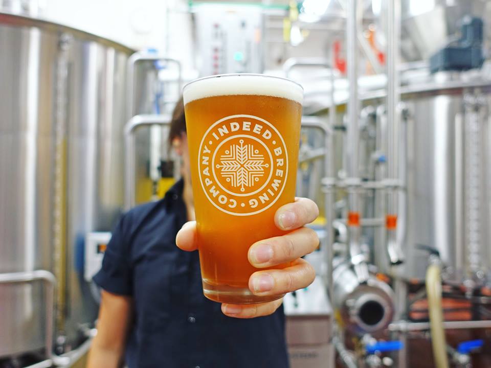 A person holding an Indeed Logo Pint Glass of beer in front of an Indeed Brewing brewery.